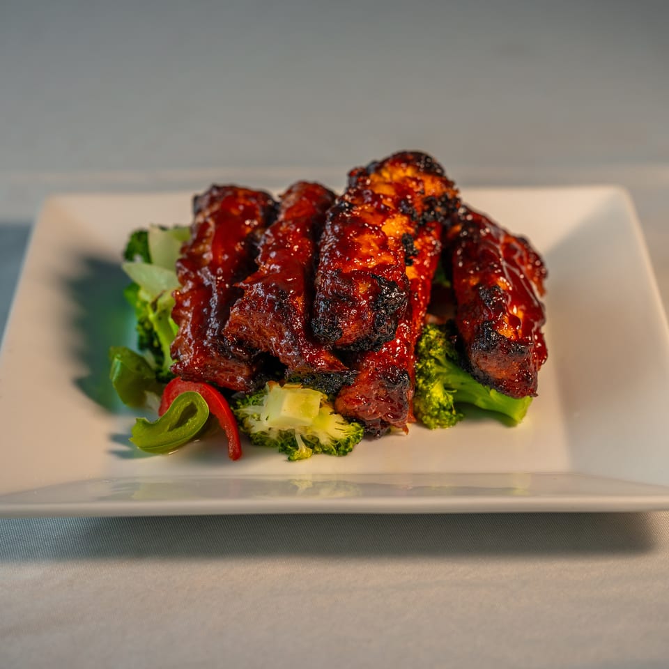 Glazed, charred grilled meat piled over broccoli, lettuce, and sliced peppers on a white rectangular plate.
