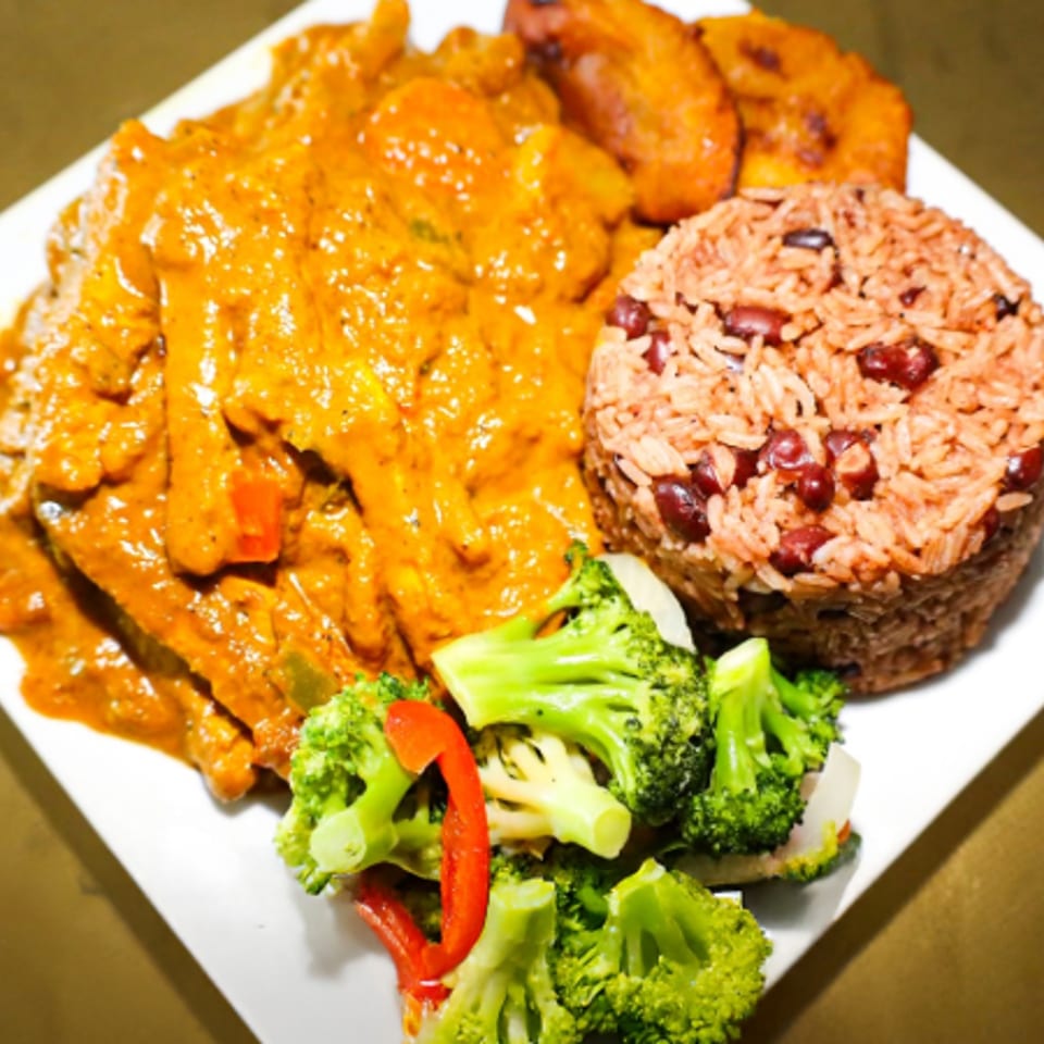 Plate of saucy chicken, rice and beans, fried plantains, and steamed broccoli with red pepper strips.
