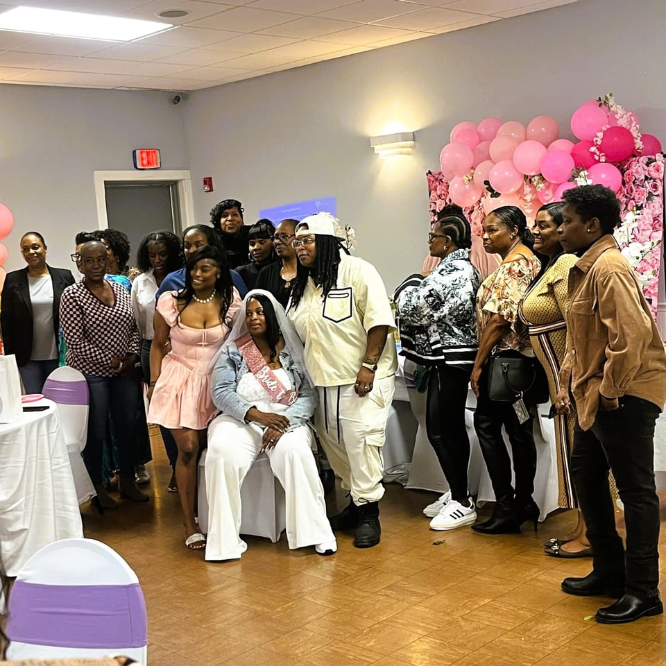 A group of women celebrating a bridal shower, with the bride seated in front of a pink balloon arch.