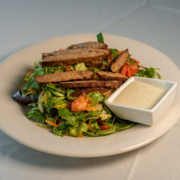 A Caribbean salad with sliced meat, mixed greens, and a side of creamy dressing on a white plate.
