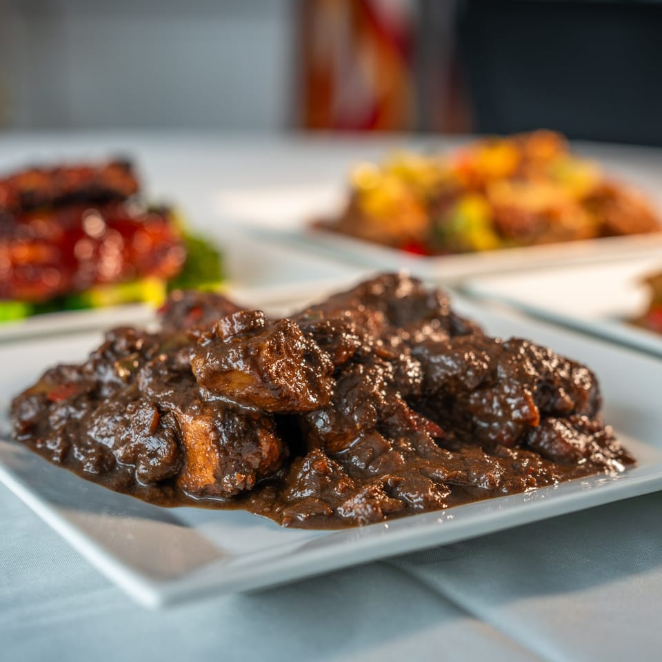 Close-up of a Caribbean stew with dark, rich sauce and meat chunks on a white plate.
