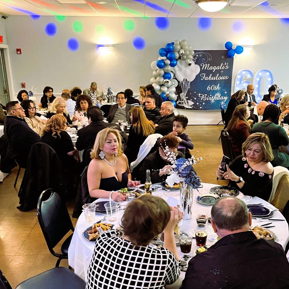 Guests seated at round tables with food and drinks, celebrating a 60th birthday with blue and silver decorations.