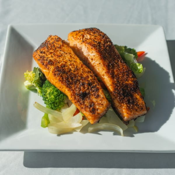Two seasoned salmon fillets on a bed of broccoli, onions, and bell peppers on a white plate.