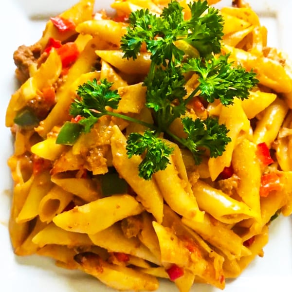 Close-up of Caribbean penne pasta with red and green peppers, meat, and parsley on a white plate.